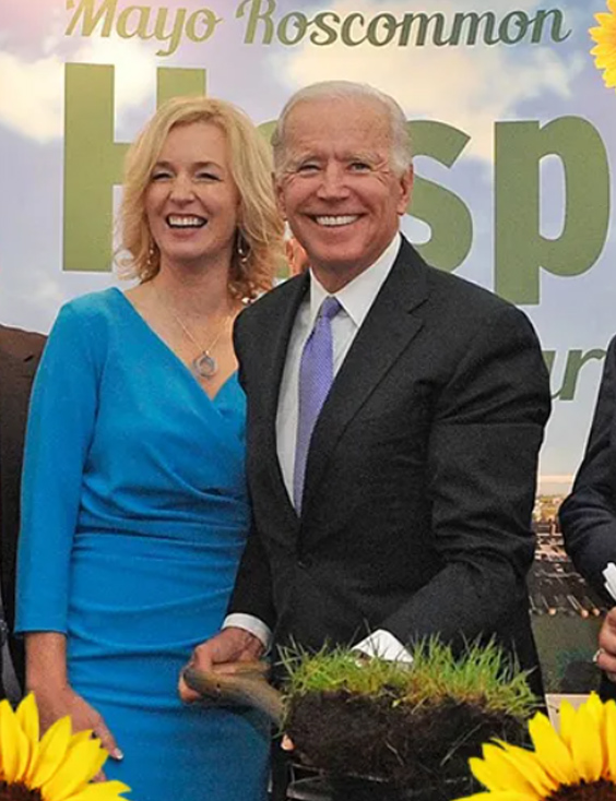 Two people smile whilst holding a patch of grass with a small spade at an event for Mayo Roscommon Hospice, with sunflowers in the foreground and a sign reading Mayo Roscommon Hospice in the background.