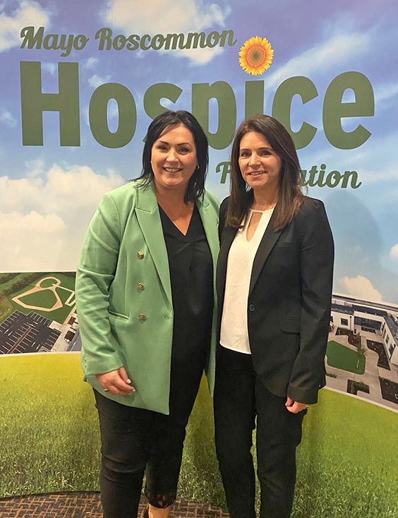 Two women in blazers stand smiling in front of a Mayo Roscommon Hospice Foundation backdrop, showcasing the organisation’s signature sunny sky and grassy field design.