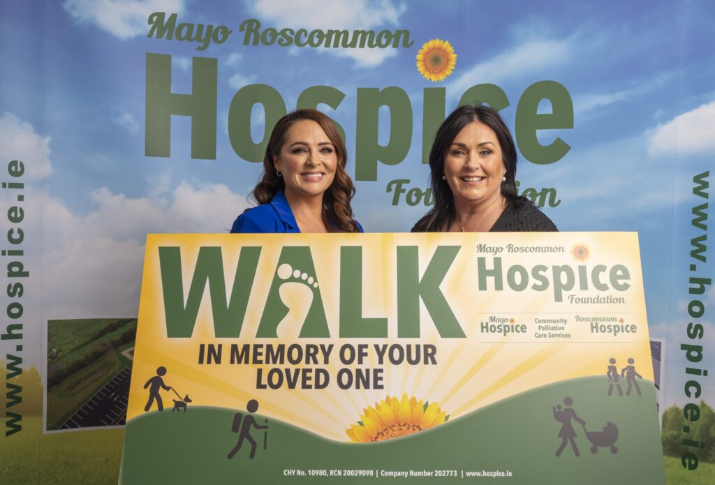 Two women smile whilst holding a sign that says WALK IN MEMORY OF YOUR LOVED ONE at a Mayo Roscommon Hospice event. The background features the Mayo Roscommon Hospice Foundation’s logo and website.