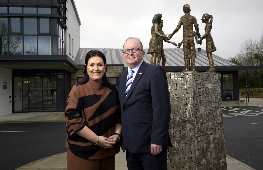 A woman and a man in business attire stand smiling in front of a modern building and the distinctive Mayo Roscommon Hospice sculpture of four children holding hands, outside on a cloudy day.