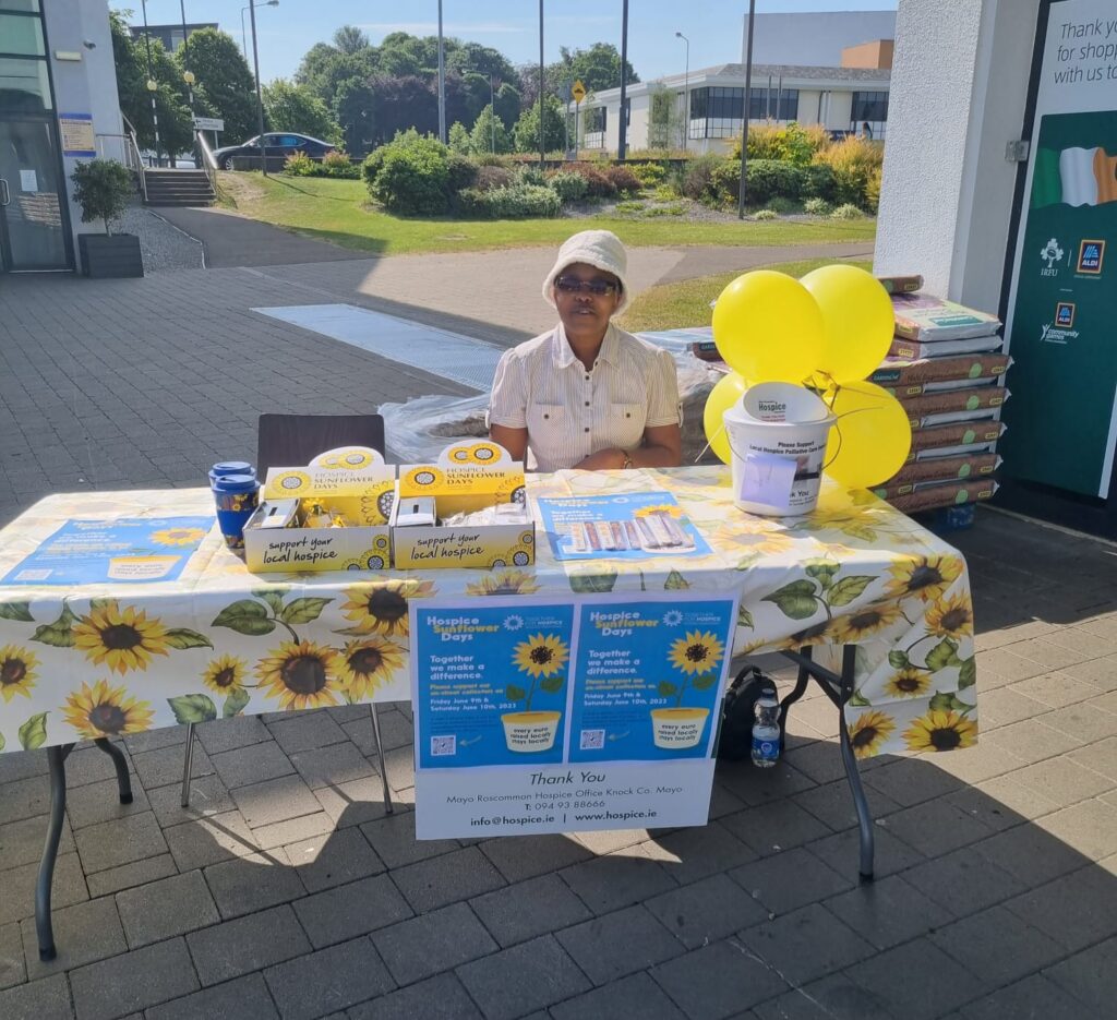 A person sits at a table decorated with sunflower-themed items, informational leaflets, donation boxes, and yellow balloons, promoting and raising funds for Mayo Roscommon Hospice outside a building on a sunny day.
