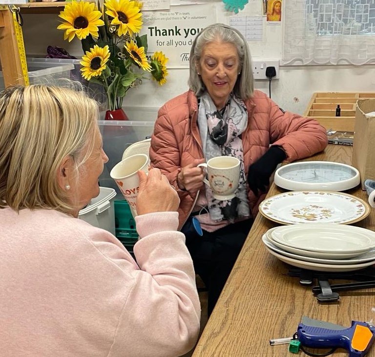 Two women sit at a wooden table in a shop, holding mugs and chatting. Amidst stacked plates, a clock, sunflowers, and various items around them, the welcoming atmosphere feels reminiscent of a Mayo Roscommon Hospice coffee morning. Both women appear relaxed and engaged in conversation.
