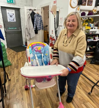 A woman smiles whilst standing behind a pink and white highchair in a room with clothes, shelves, and wooden flooring, showing her support for Mayo Roscommon Hospice.