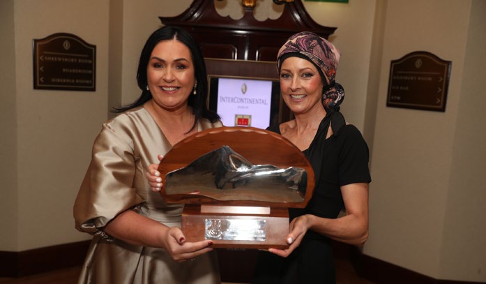 Two women smiling and holding a large, shiny Mayo Roscommon Hospice award with a mountain design on it, standing indoors in formal attire.