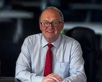 A man wearing glasses, a white shirt, and a red tie sits at a desk, smiling at the camera in an office setting, proudly displaying his support for Mayo Roscommon Hospice.
