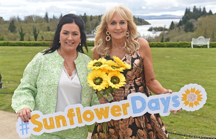 Two women standing outdoors, one holding sunflowers, both smiling and holding a sign that reads #SunflowerDays with a garden and water in the background, celebrating Mayo Roscommon Hospice’s annual event.