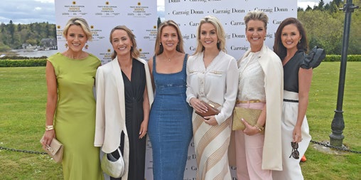 Six women dressed in stylish spring outfits pose and smile together outdoors in front of a branded Mayo Roscommon Hospice backdrop on a grassy lawn.