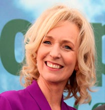 A smiling woman with shoulder-length blonde hair wearing a purple blazer stands in front of a blue and green background, representing Mayo Roscommon Hospice.