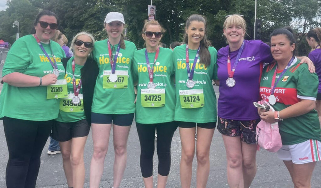 Seven women stand together outdoors, smiling after a race, proudly wearing medals. Five wear matching green Mayo Roscommon Hospice shirts, one wears a purple shirt, and another sports a Mayo jersey. Trees and other participants can be seen in the background.
