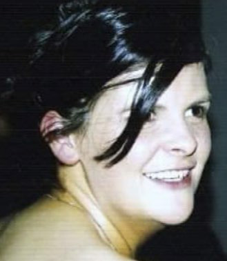 A woman with dark hair styled up smiles whilst looking slightly to the side. She is in a dimly lit setting, and her shoulder is visible in the foreground, capturing a moment of quiet reflection at a Mayo Roscommon Hospice event.