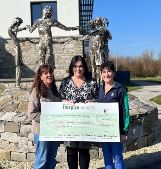 Three women stand outside, smiling and holding a large cheque for €1,610 made out to the Mayo Roscommon Hospice. In the background, a metal sculpture of people holding hands emphasises the spirit of community and support for the Mayo Roscommon Hospice Foundation.