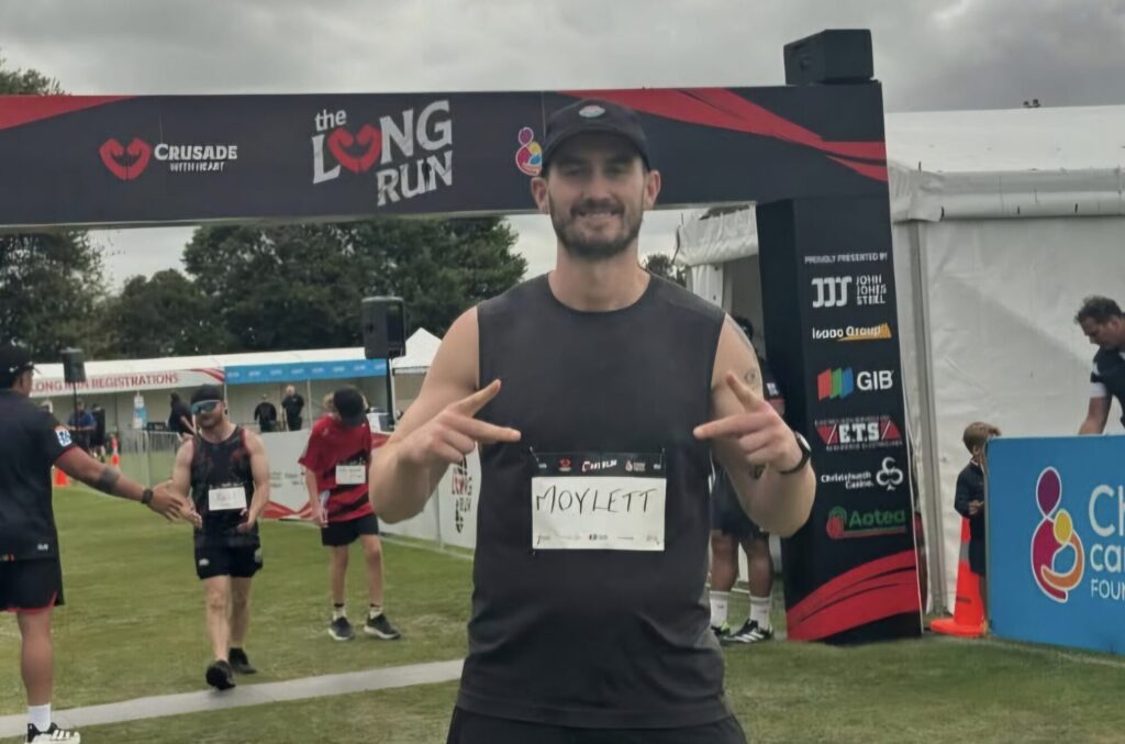 A runner stands smiling and making peace signs at a Mayo Roscommon Hospice race event, wearing a name tag labelled Moylett, with other participants and an event banner visible in the background.