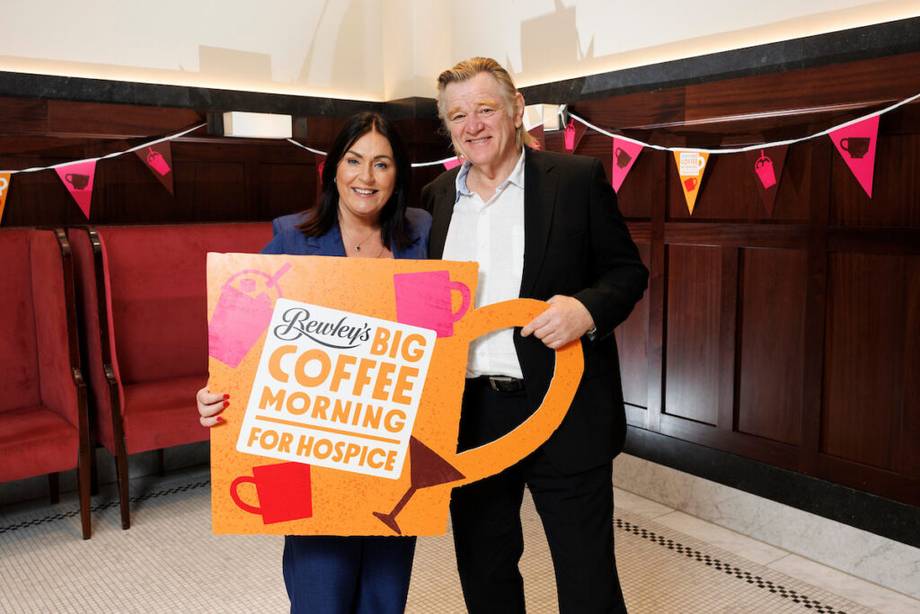 Two people smile while holding a large sign that reads, Bewleys Big Coffee Morning for Hospice, showing their support for Mayo Roscommon Hospice. Pink bunting decorates the background.