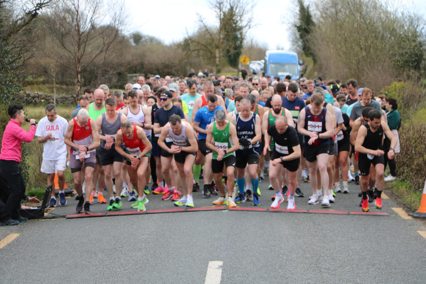 A group of runners in sports kit are lined up at the starting line of a road race, preparing to begin. Spectators and trees line the road in the background, as participants gather to support Mayo Roscommon Hospice.
