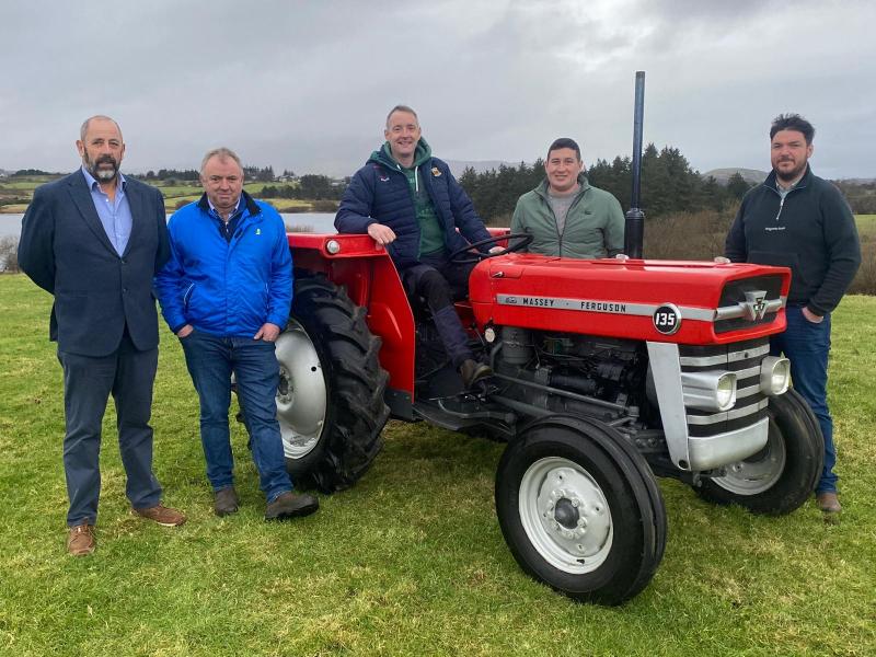 Five men stand outdoors on grass beside a red Massey Ferguson 135 tractor, with trees and water in the background under a cloudy sky, proudly showing their support for Mayo Roscommon Hospice.