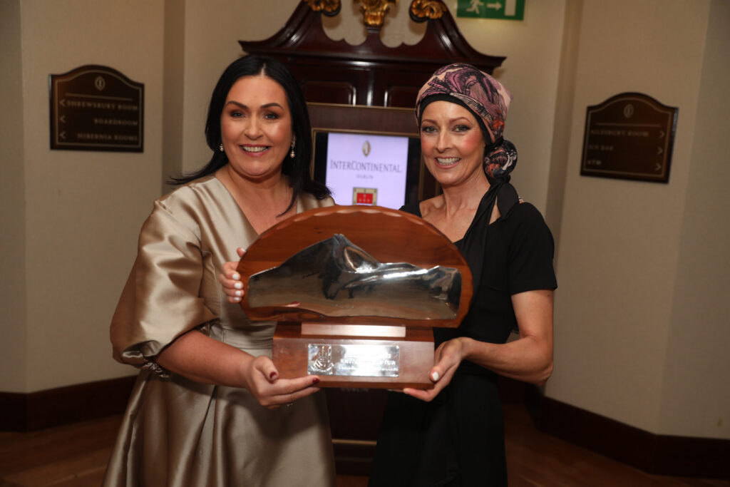 Two women smiling and holding a trophy together in an indoor setting, with a sign for Mayo Roscommon Hospice and wooden plaques visible in the background. One woman is wearing a headscarf.