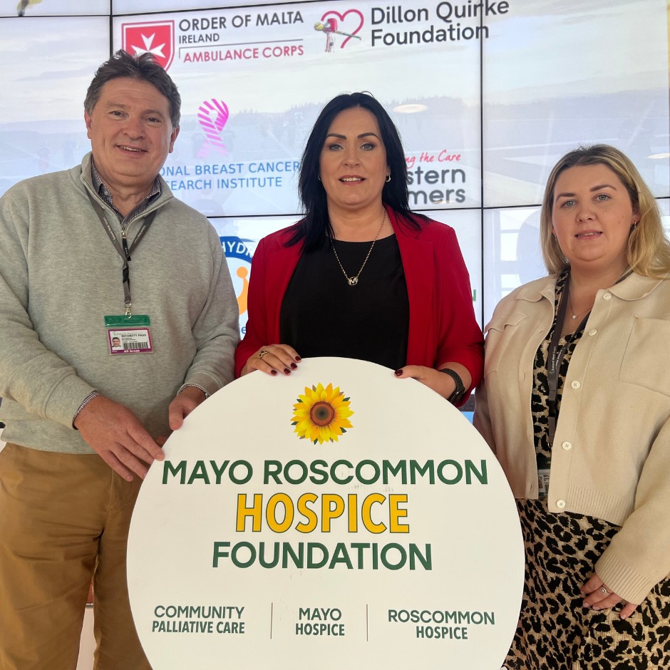 Three people stand holding a Mayo Roscommon Hospice Foundation sign, proudly supporting the invaluable work of Mayo Roscommon Hospice. Logos for charities and organisations are displayed on a screen behind them.