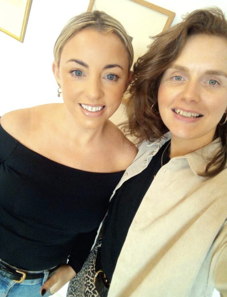 Two women smiling and posing for a selfie indoors in support of Mayo Roscommon Hospice; one wears a black off-shoulder top, and the other a beige shirt with a black top underneath.