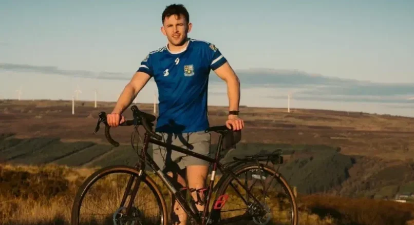 A man in a blue sports shirt stands outdoors holding a bicycle, with hills and wind turbines visible in the background under a clear sky, supporting the Mayo Roscommon Hospice.