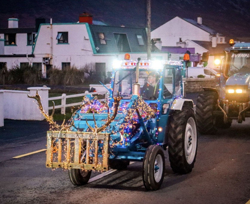 A blue tractor decorated with lights and fake antlers drives down a street in support of the Mayo Roscommon Hospice, followed by another tractor, with houses and a mountain in the background.