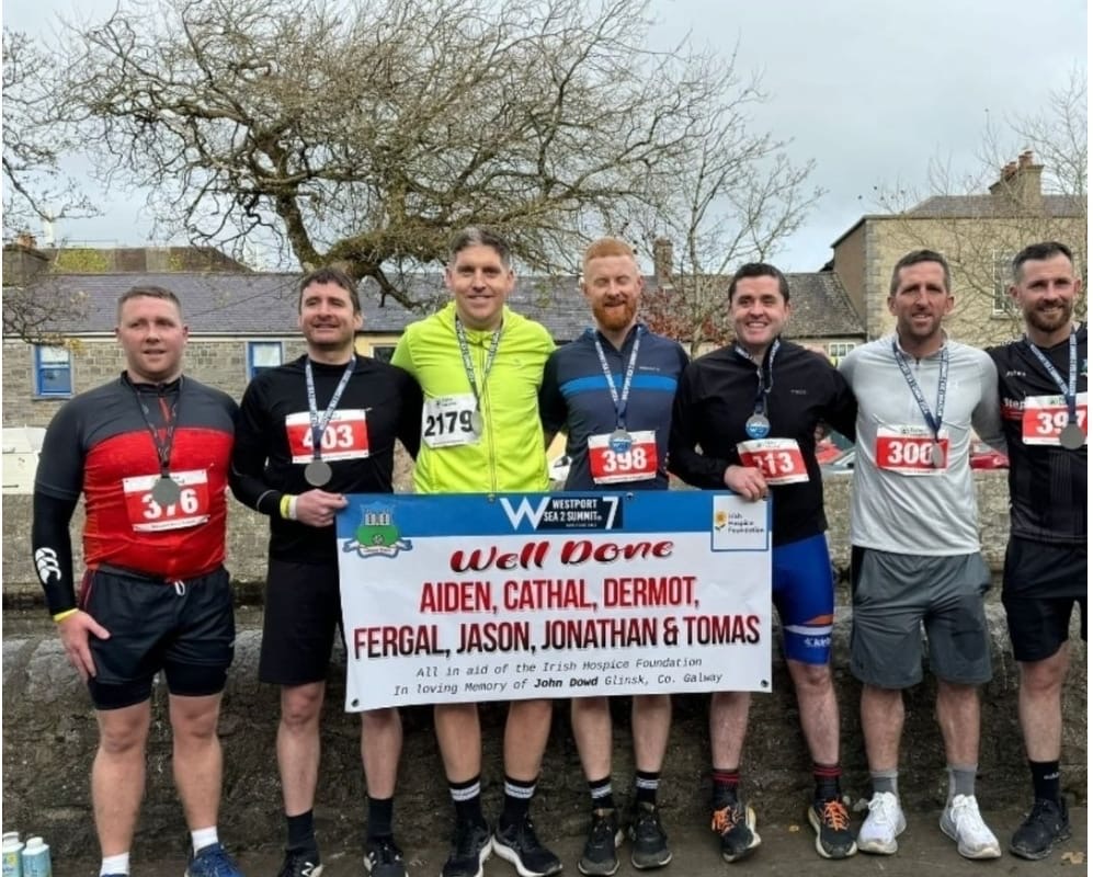 Seven men in sportswear stand outdoors, smiling and holding a Well Done banner with their names and medals after completing a race in support of Mayo Roscommon Hospice. Houses and leafless trees are visible in the background.