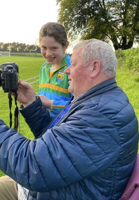 An older man in a blue jacket shows a digital camera to a smiling girl in a colourful sports jersey. They are sitting outdoors on the grass near a sports pitch, with trees in the background, during an event supporting Mayo Roscommon Hospice.