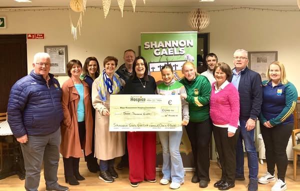 A group of people pose indoors with a large donation cheque for €3,770, made out to Mayo Roscommon Hospice, in front of a Shannon Gaels banner. Some wear green and yellow sports jerseys.