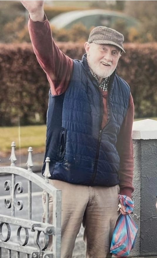 An older man wearing a flat cap, blue waistcoat, and red jumper smiles and waves whilst standing by a metal gate, holding a colourful bag in his left hand—perhaps on his way to support the Mayo Roscommon Hospice. Bushes and a lawn are visible in the background.