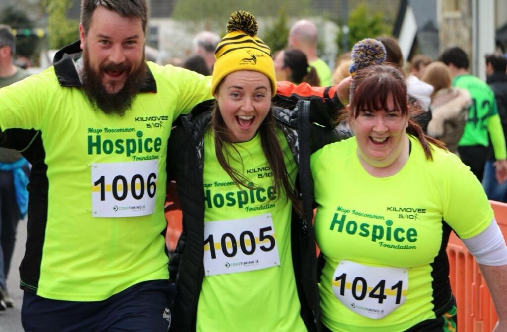 Three enthusiastic people wearing bright yellow Mayo Roscommon Hospice shirts and race numbers (1006, 1005, 1041) smile with arms round each other at an outdoor event, with other participants visible in the background.