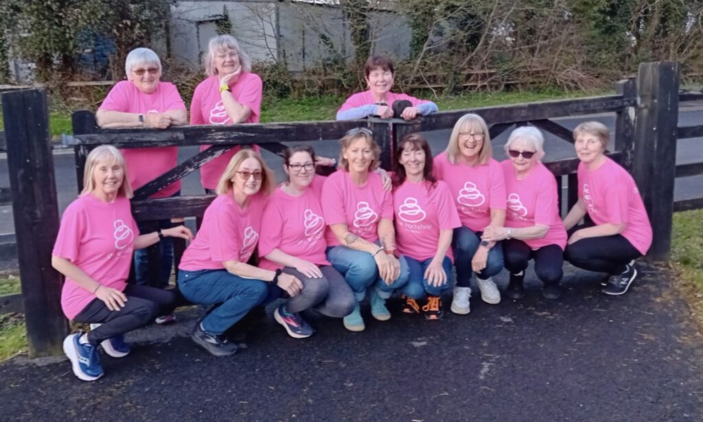 Eleven women pose outdoors in front of a wooden gate, all wearing matching bright pink T-shirts in support of Mayo Roscommon Hospice. The group is smiling, and trees and a building are visible in the background.