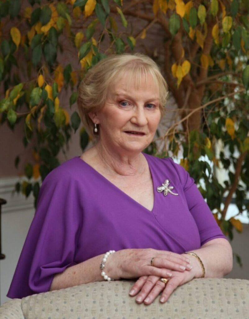 An older woman with short blonde hair, wearing a purple dress with a brooch and jewellery, sits smiling on a beige chair. Green and yellow leaves fill the background, creating a warm atmosphere reminiscent of the comforting care provided by Mayo Roscommon Hospice.