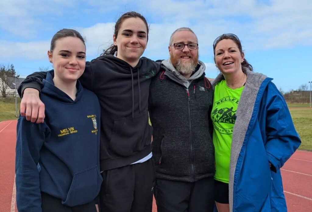 Four people stand arm in arm on an athletics track, smiling at the camera; two are teenagers, and two are adults. The sky is partly cloudy, and everyone is dressed in casual sportswear, proudly supporting Mayo Roscommon Hospice at this community event.