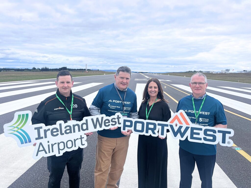 Four people stand on an airport runway, holding Ireland West Airport and Portwest signs in support of Mayo Roscommon Hospice. They wear branded shirts and lanyards. The sky is cloudy above them and runway lines are visible beneath their feet.