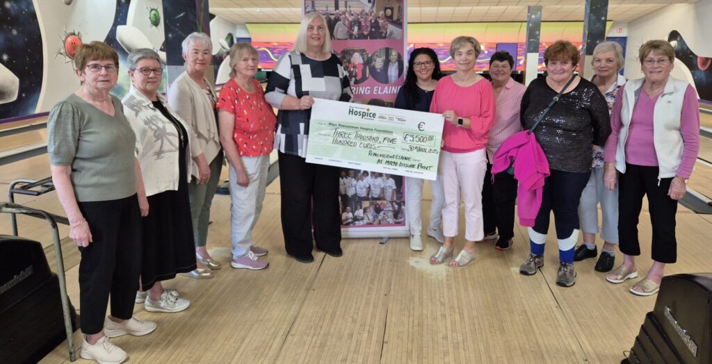 A group of women, standing on a bowling alley, present a large cheque for €500 to the Mayo Roscommon Hospice charity. They smile and pose together, with a Caring Elaine banner in the background.