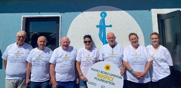 Seven people wearing white T-shirts with Mayo Roscommon Hospice text stand in front of a blue wall with an anchor symbol, holding a Mayo Roscommon Hospice Foundation sign under a bright, sunny sky.