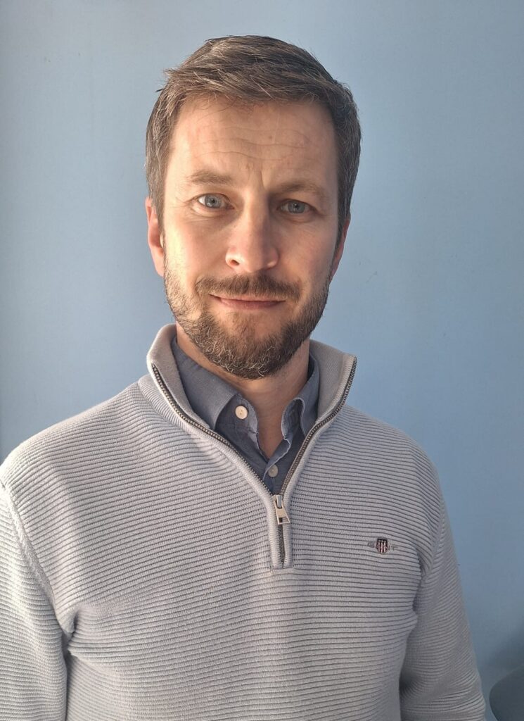 A man with short brown hair and a beard is wearing a light grey zip-up jumper over a blue collared shirt, standing in front of a plain blue background, looking at the camera with a slight smile. He appears to be supporting the Mayo Roscommon Hospice, as indicated by the subtle logo on his jumper.
