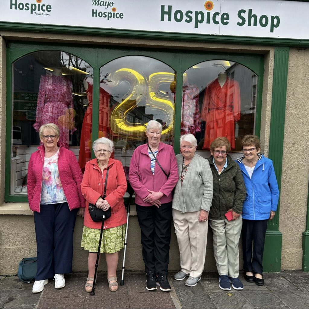 Six elderly women stand smiling in front of the Mayo Roscommon Hospice Shop with large gold 25 balloons and dresses in the window behind them. The shop is painted green with white signage above.