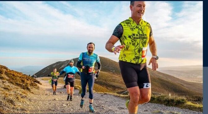 Four runners in athletic kit smile as they ascend a rocky mountain trail under a blue sky, with hills and valleys visible in the background, proudly supporting Mayo Roscommon Hospice on their challenging journey.