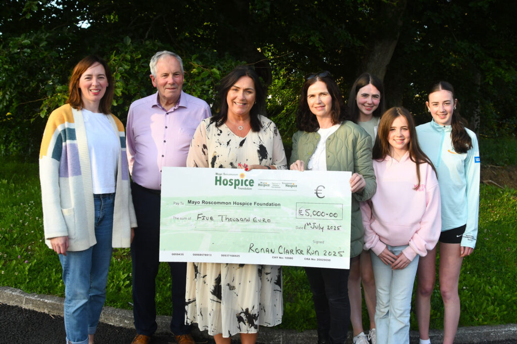 Six people stand outdoors, smiling and holding a large donation cheque for €5,000 from the Ronan Clarke Run 2023 to the Mayo Roscommon Hospice. Trees and grass are visible in the background.