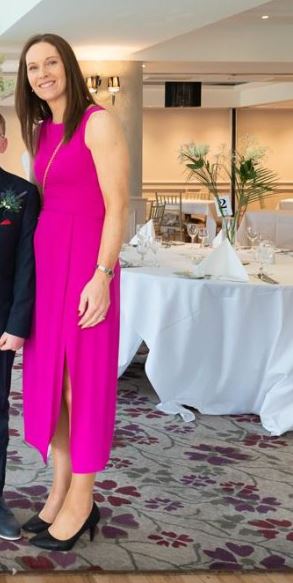 A woman in a bright pink sleeveless dress and black heels stands indoors beside a set dining table with white linen and floral arrangements, attending an event in support of Mayo Roscommon Hospice.