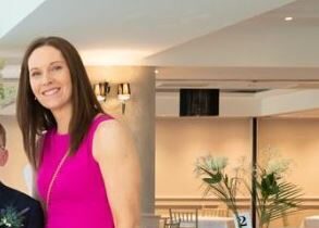 A woman with straight brown hair, wearing a sleeveless bright pink dress, stands indoors near some decorative plants and tables in a well-lit room, showing her support for the Mayo Roscommon Hospice.