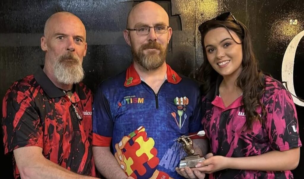 Three people pose together, two men and one woman, all wearing colourful shirts. The man in the centre holds a trophy featuring a puzzle piece design, proudly recognising their contribution to Mayo Roscommon Hospice. They are standing against a dark background.