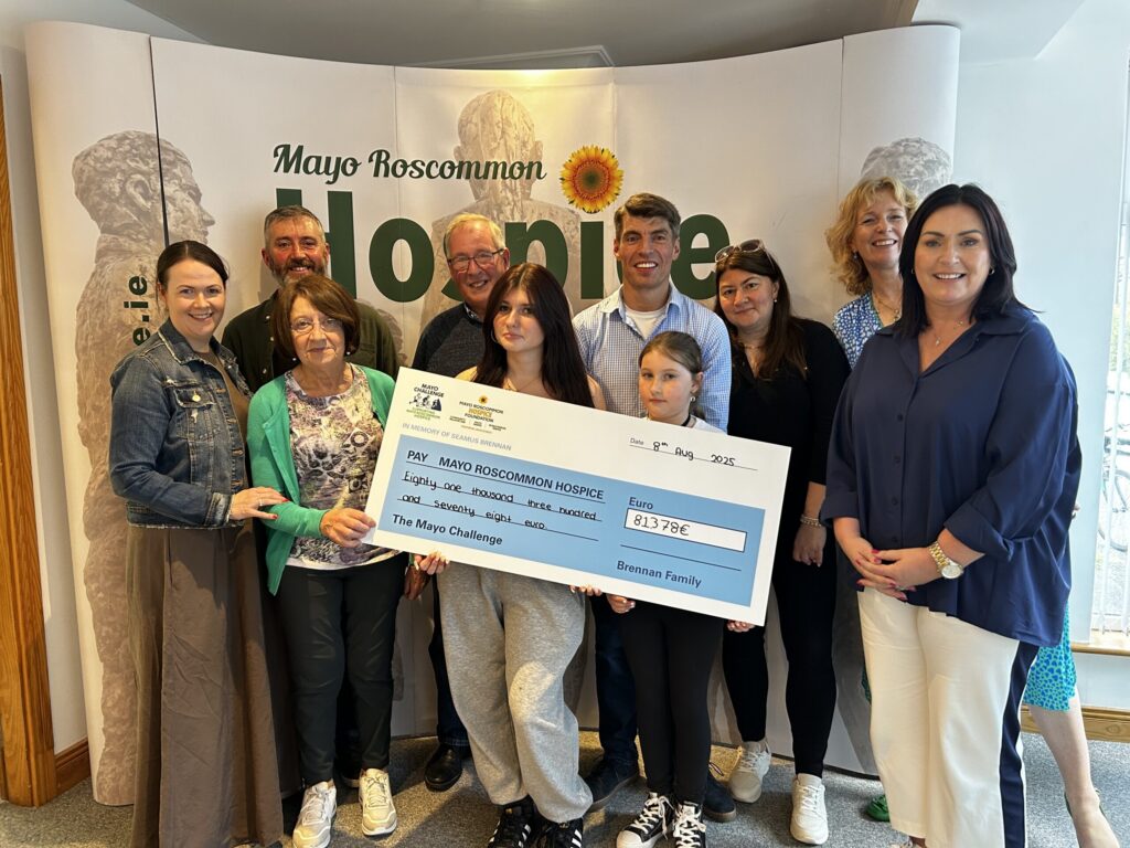 Nine people stand together holding a large charity cheque for Mayo Roscommon Hospice. They are smiling, and the background features the Mayo Roscommon Hospice logo and a sunflower image.