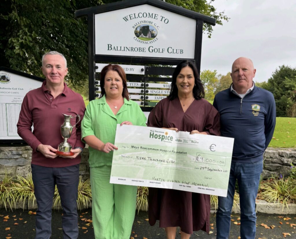 Four people stand outside Ballinrobe Golf Club. One holds a trophy and two hold a large cheque for €9,000 to Mayo Roscommon Hospice Foundation, proudly supporting Mayo Roscommon Hospice. There is a golf club sign and green trees in the background.