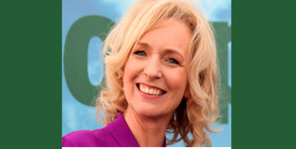 A smiling woman with blonde, wavy hair wearing a purple top stands in front of a blue and green background, representing the compassionate care provided by Mayo Roscommon Hospice.