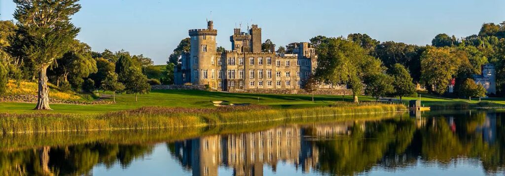 A stone castle sits beside a calm lake, surrounded by green grass and trees, with its reflection visible in the water under a clear blue sky—a tranquil scene reminiscent of the peaceful surroundings near the Mayo Roscommon Hospice.