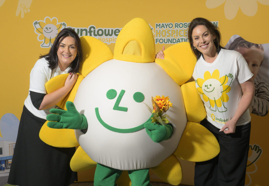 Two women in sunflower-themed shirts stand beside a person in a sunflower mascot costume holding flowers, all smiling in front of a yellow Mayo Roscommon Hospice Sunflower Foundation backdrop.