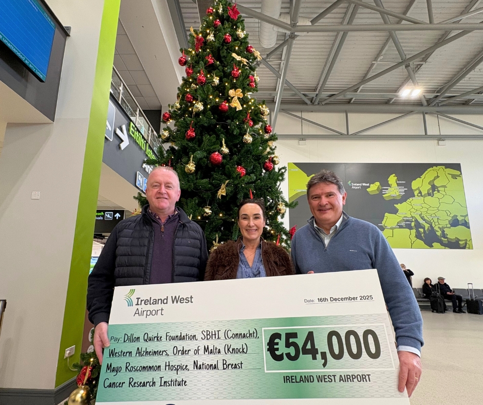 Three people stand in front of a Christmas tree at an airport, holding a large €54,000 charity cheque for Mayo Roscommon Hospice. A map of Ireland is visible in the background, along with airport signage and seating.