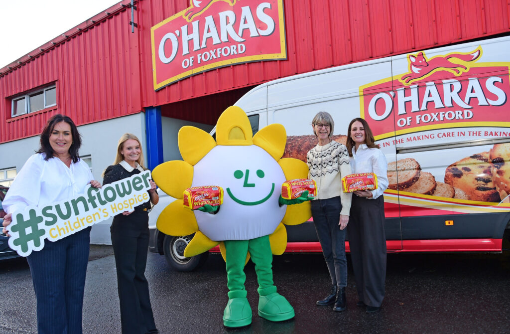 Five people, including one in a sunflower mascot costume, stand smiling in front of an O’Haras of Foxford van, holding Mayo Roscommon Hospice and food signs and packages.