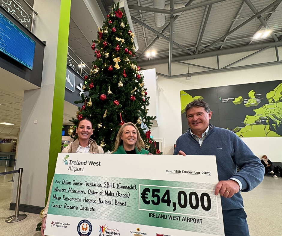 Three people hold a large €54,000 cheque in front of a decorated Christmas tree inside Ireland West Airport, celebrating a generous donation to Mayo Roscommon Hospice. A map of Europe is visible on the wall behind them.